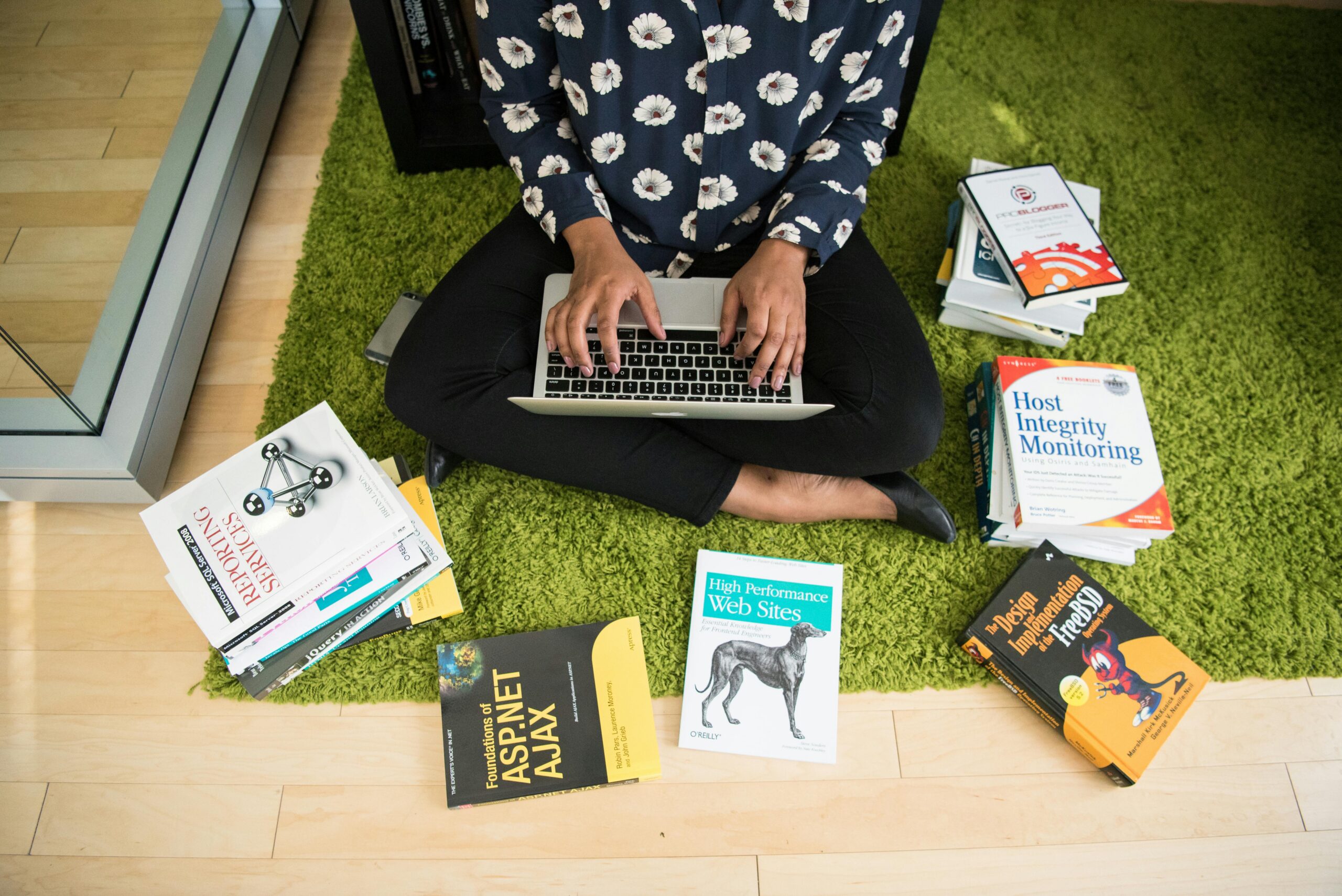 Yapay Zeka Haberleri: Geleceği Şekillendiren Gelişmeleri Yakından Takip Edin Woman sitting on green rug working on laptop, surrounded by technology books in a modern room.